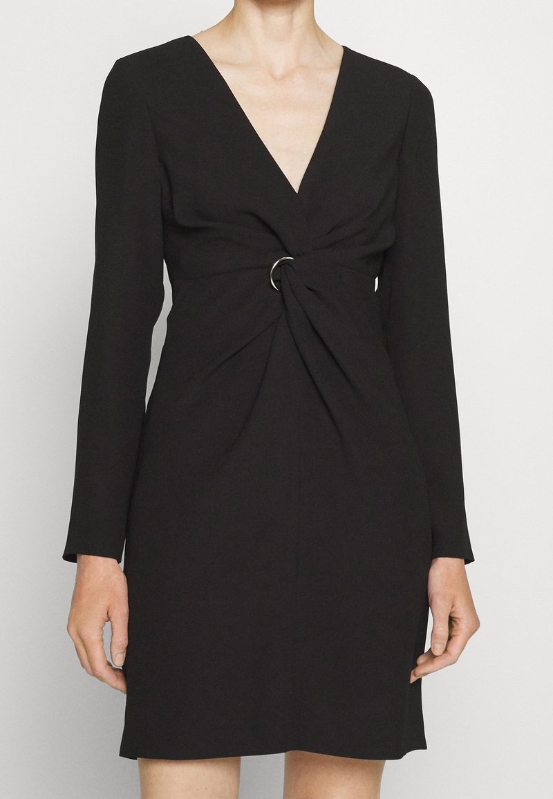 Femme portant une robe noire à manches longues avec un col en V profond et un détail d'anneau argenté réunissant le tissu à la taille.