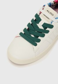 White leather sneaker with green laces, rounded toe, and a patterned inner lining. Features a branding tag on the tongue.