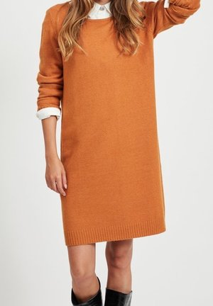 Kobieta ubrana w pomarańczową sukienkę z długim rękawem na sweter, nałożoną na białą koszulę z kołnierzykiem, w połączeniu z czarnymi kozakami za kolano, na jednolitym tle.