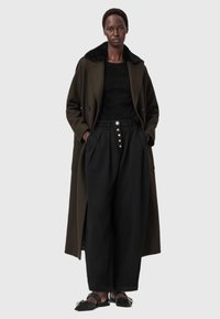 Casaco longo verde-oliva com colarinho de pelagem preta, combinado com calças pretas de cintura alta com detalhes em botão e sapatos planos pretos.