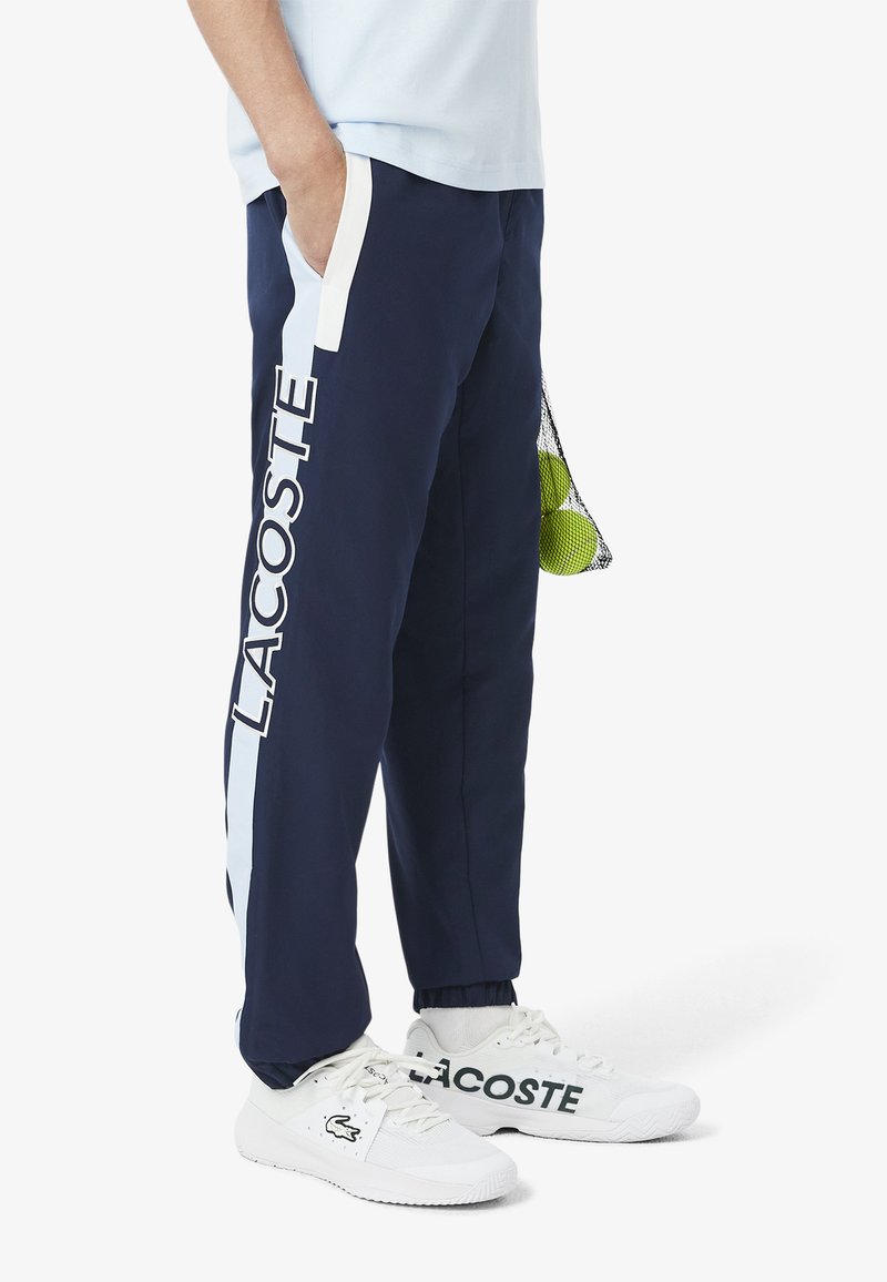 Donkerblauwe joggers met witte accenten aan de zijkant en het "LACOSTE" logo. Gestyled met witte sneakers met logo aan de zijkant.