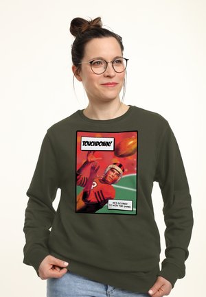 Kvinde iført briller og en olivengrøn sweatshirt med en tegneseriestil fodboldspiller, der griber en bold, og teksten "TOUCHDOWN! Han har scoret for at vinde kampen!"