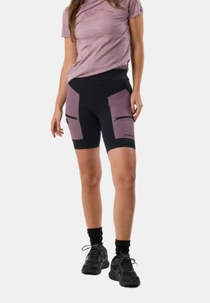 Femme portant un short de cyclisme noir et mauve avec des poches zippées, un t-shirt mauve, des chaussettes noires et des chaussures de sport noires, debout sur un fond blanc.