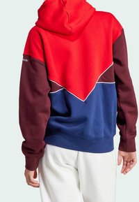 Mikina s kapucí v červené, bordó a modré barvě. Vyznačuje se designem ve stylu bloků barev s kontrastními panely a bílým lemováním podél švů.