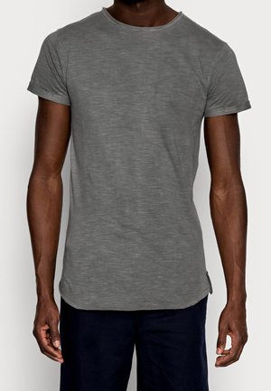 Homme portant un T-shirt gris à manches courtes ajusté et un pantalon sombre, debout contre un fond clair, les bras détendus le long du corps.