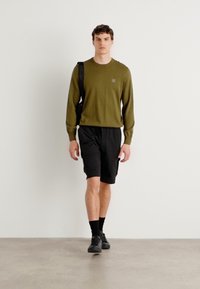 Pulover lung cu mâneci, de culoare verde măslin, pantaloni scurți negri și pantofi negri. Poartă o geantă de umăr neagră, stând pe un fundal neutru.
