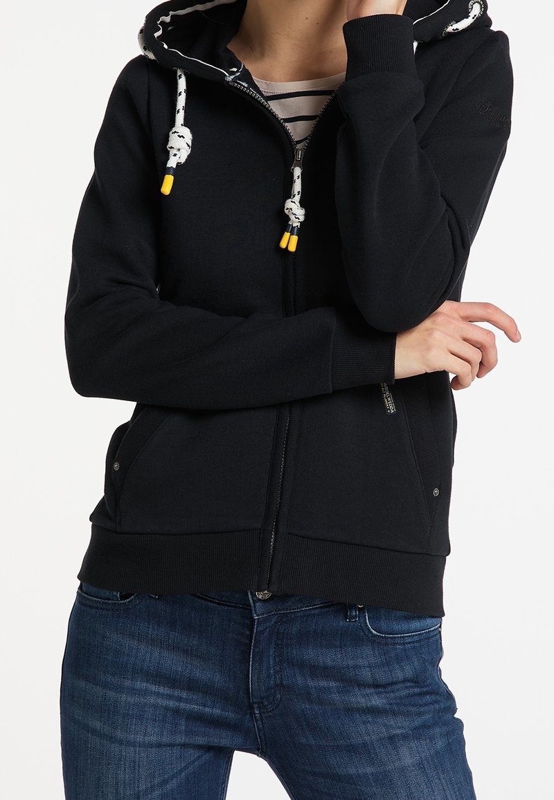 Personne portant un sweat à capuche noir zippé avec des cordons blancs et des embouts jaunes, associé à un jean bleu et une chemise rayée en dessous.