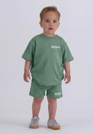 Toddler standing and wearing green oversized t-shirt and matching shorts with white sneakers on a plain background.