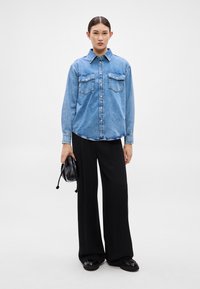 Džínová košeľa svetlomodrej farby s gombíkmi vpredu, dvomi náprsníkovými vreckami a klasickým golierom, v kombinácii s čiernymi nohavicami s širokými nohavicami a čiernou taškou.