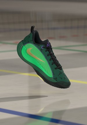 Зелена и черна спортна обувка със златисто лого swoosh, плаваща над пода на спортна зала с цветни линии на игрището.