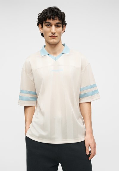 Jeune homme aux cheveux bouclés et foncés, portant une chemise ample crème avec un col et des rayures sur les manches bleu pâle, debout devant un fond clair uni.