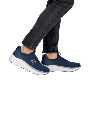 Personne portant des baskets slip-on bleu marine avec semelles blanches et jean noir, un pied légèrement levé sur fond blanc.