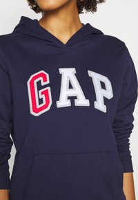 Felpa navy in cotone con tasca a marsupio. Presenta il logo ricamato "GAP" in rosso, bianco e azzurro sul davanti.