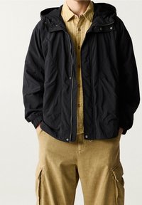 Giacca nera leggera con cappuccio, dotata di zip frontale e tasche laterali, indossata sopra una camicia a quadri gialla e pantaloni cargo color khaki.