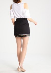 Czarna spódnica z kolorowym, haftowanym wykończeniem w kwiaty, zestawiona z białą bluzką z odkrytymi ramionami. Błyszczące beżowe buty na słupku dopełniają stylizację.