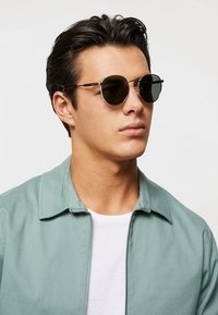 Ochelari rotunzi cu lentile întunecate, rame subțiri din metal și accente discrete. Purtați de o persoană îmbrăcată într-o jachetă verde deschis peste o cămașă albă.