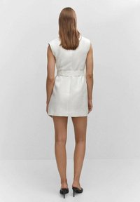 Biała lniana sukienka bez rękawów z dopasowaną talią i detalem w postaci paska, o prostym kroju i minimalistycznym designie. Czarne szpilki.