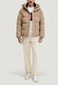 Beige vadderad jacka med dragkedja, stora quiltade sektioner och huva. Bärs över en vit sweatshirt med rund halsringning och beige byxor.