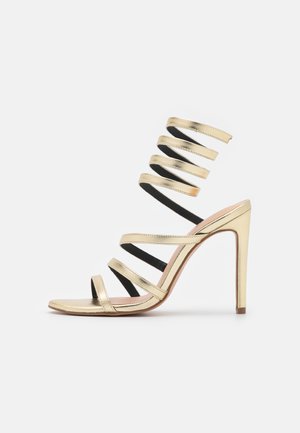 Sándal de tacón alto dorado con múltiples tiras finas, incluyendo una tira en espiral que envuelve el tobillo, exhibido sobre un fondo blanco.