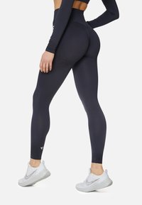 Woman wearing black high-waisted leggings and white Nike sneakers, posing with one hand on hip and the other relaxed by side.