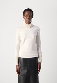 Svijetlo kremasti dugi rukavi majica s rebrastim turtleneck-om, koja ima ukrasnu dugmad. Uparen s crnom kožnom suknjom.