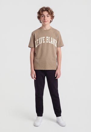 Adolescent debout face à l’avant portant un t-shirt marron avec le texte "LEAVE BLANK", un pantalon noir et des baskets blanches sur un fond clair uni.