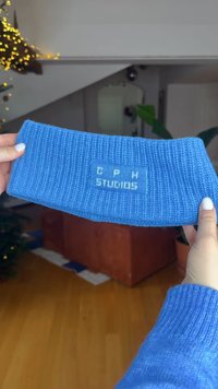 Blauwe geribbelde gebreide hoofdband met een rechthoekige patch met de tekst "CPH Studios" in contrasterend lichtblauw. Zachte textuur, lichtgewicht ontwerp.
