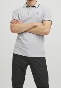 Ljusgrå polo skjorta med svart krage och detaljer, korta ärmar och plagg med tre knappar. Bärs med mörka byxor, armarna i kors.