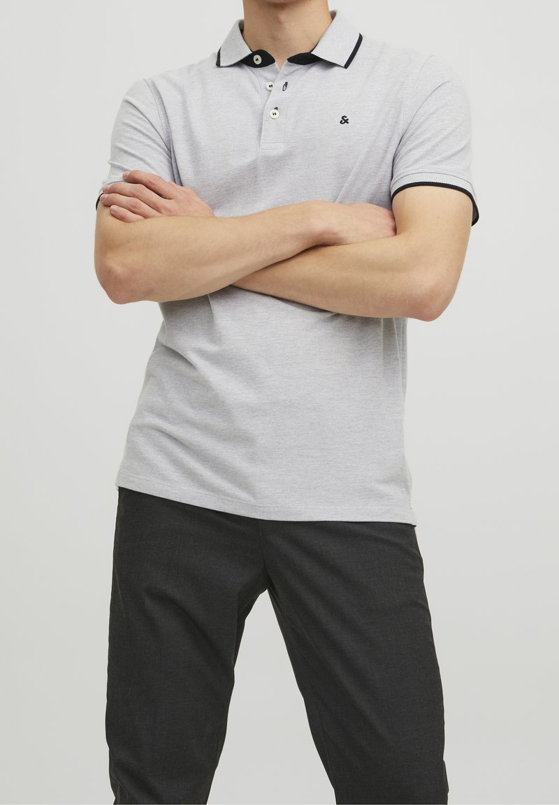 Ljusgrå polo skjorta med svart krage och detaljer, korta ärmar och plagg med tre knappar. Bärs med mörka byxor, armarna i kors.