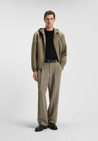Lichte, olijfgroene jas met een capuchon, gecombineerd met een zwart shirt en beige wijde broek. Gladde structuur, met zwarte schoenen.