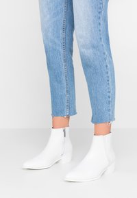 Botas de tobillo de cuero blanco con punta afilada y tacón bajo en bloque, con un zipper lateral para facilitar su uso. Combinadas con jeans de un azul claro desgastados.