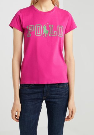 Kobieta ubrana w jaskraworóżowy t-shirt z krótkim rękawem, z zielonym napisem "POLO" i logo gracza polo na koniu, w połączeniu z ciemnoniebieskimi dżinsami.