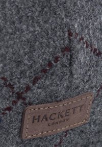 Braunes Lederetikett von Hackett London, auf grauem strukturiertem Stoff mit feinem roten Karo-Muster genäht.