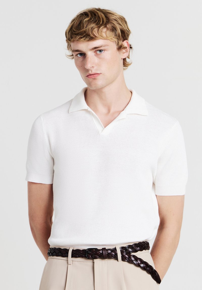Polo shirt bianco lavorato a maglia con colletto e maniche corte, caratterizzato da un tessuto testurizzato. Abbinato a pantaloni beige chiaro e una cintura in pelle intrecciata.