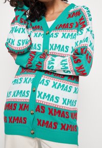 Turkusowy kardigan z powtarzającym się czerwonym i zielonym napisem "XMAS". Wzór dzianinowy, zapięcia na guziki, ściągacze przy mankietach oraz kontrastowy dekolt.