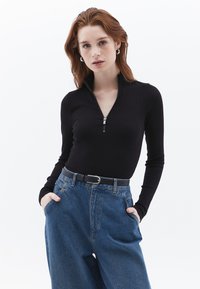 Zwarte ritssluiting met lange mouwen, gecombineerd met lichtblauwe spijkerbroek met hoge taille. De outfit bevat een zwarte riem met een zilveren gesp.