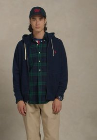 Casaco de capuz azul-marinho com fecho em cima de uma camisa às riscas em verde escuro e azul, e calças bege; usa um boné preto com letras vermelhas e um relógio.