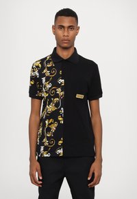 Svart og mønstret polo-skjorte med blomsterdesign i gul og hvit på den ene siden, med lomme og logo-merke på brystet.