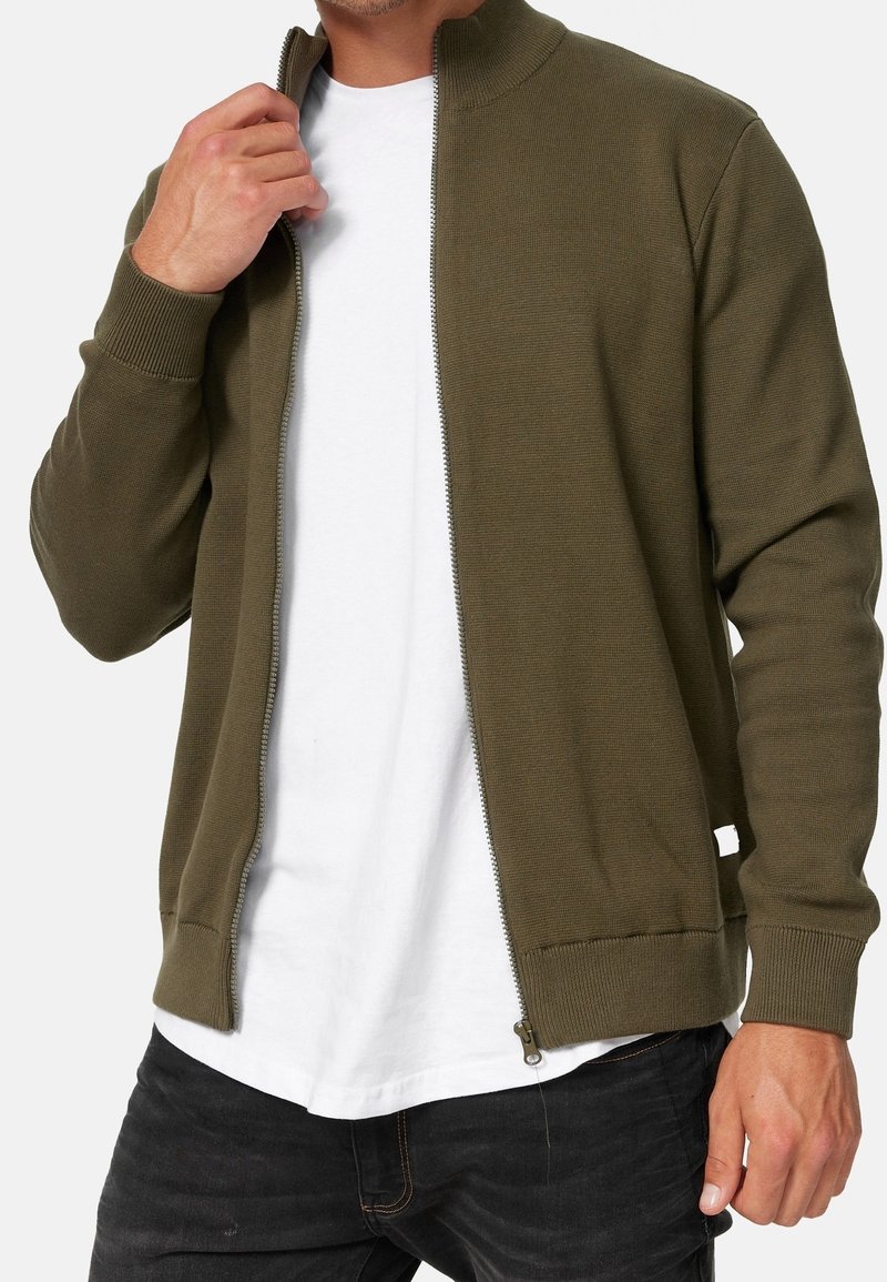 Homme portant un pull zippé vert olive sur un t-shirt blanc et un jean noir, main tenant le pull près du col.