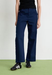 Personne portant un pantalon large bleu marine, un top sans manches noir et des baskets noires avec des rayures blanches, se tenant sur un sol vert.