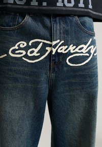 Close-up of blue denim jeans with white cursive "Ed Hardy" logo across the front waistband and pocket area.