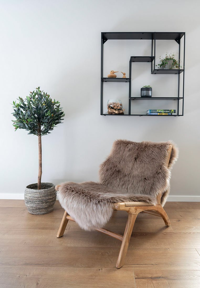 Chaise en bois avec un coussin et un dossier en fourrure brun clair, accompagnée d'un pot en gris texturé et d'un arbre artificiel, contre un mur neutre.