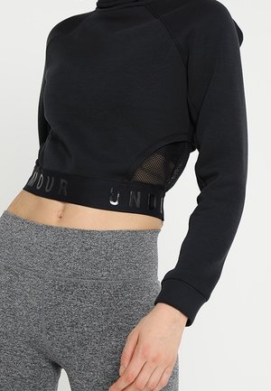 Frau trägt ein schwarzes tailliertes Sporttop mit Netzseitenteilen und graue, hoch taillierte Leggings, zeigt den Bauch und hat die rechte Hand auf dem Oberschenkel ruhend.