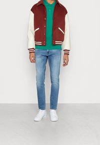 Bordowa kurtka varsity z kremowymi rękawami, zielona koszulka polo, jasnoniebieskie dżinsy i białe sneakersy. Posiada ściągacze na rękawach oraz boczne kieszenie.