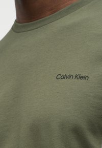 Κοντινό πλάνο σε ελιάς πράσινη μπλούζα με στρογγυλή λαιμόκοψη και μικρό μαύρο λογότυπο "Calvin Klein" στο αριστερό στήθος, που φοράει ένα άτομο.