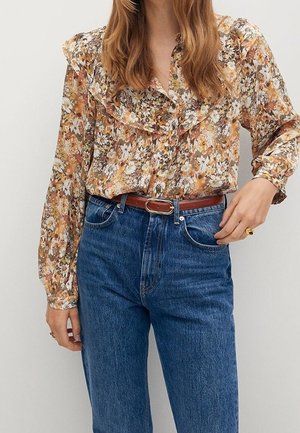 Kvinde iført en blomsterprintet bluse med lange ærmer, stoppet ned i højttaljede blå jeans med et brunt bælte, står foran en ensfarvet baggrund.