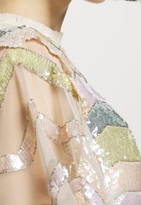 Gros plan sur une personne portant un haut transparent orné de formes géométriques à paillettes aux couleurs pastel et avec un col bordé de dentelle.