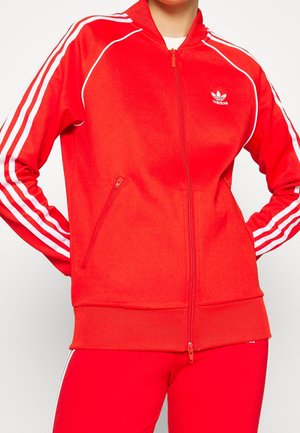 Person iført en rød Adidas træningsjakke og matchende bukser med hvide striber på ærmer og ben, stående med hænderne på hoften.