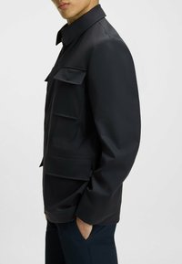 Chaqueta negra con un cuello tipo solapa, que presenta dos bolsillos en el pecho y una textura suave y mate. Diseño ajustado, con bolsillos laterales visibles.