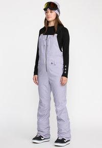 Salopette de neige lilas clair avec fermeture éclair �à l'avant, bretelles réglables et deux poches avant. Associée à un t-shirt noir à manches longues et des baskets noires.